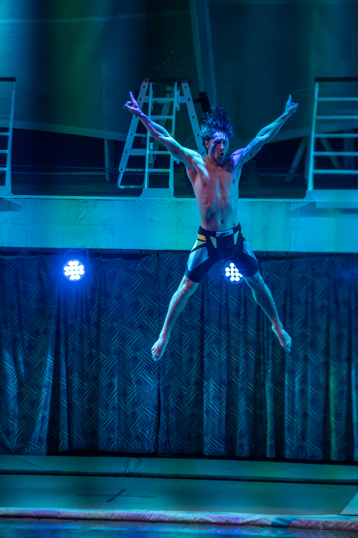 Performer midair above the water under blue stage lights
